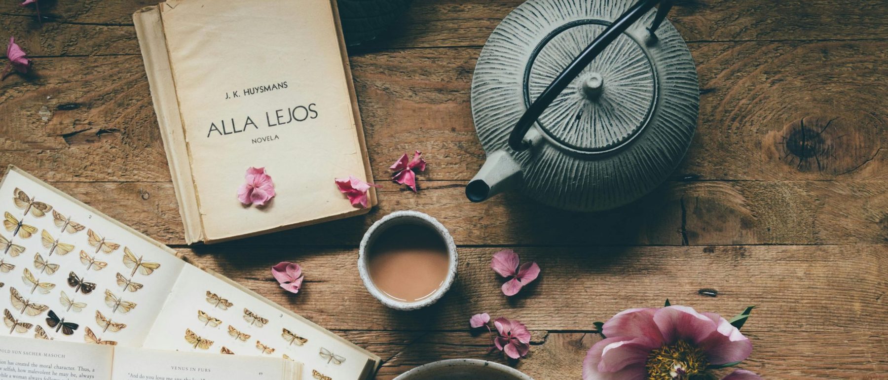 Old books, a cast-iron teapot and a cup of tea on a wooden table scattered with pink petals